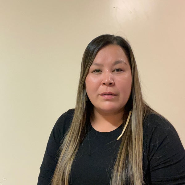 Janelle Pierrot stands in front of a beige wall wearing a black long sleeve top. Her hair goes past her shoulders and past the cropping of the photo. 