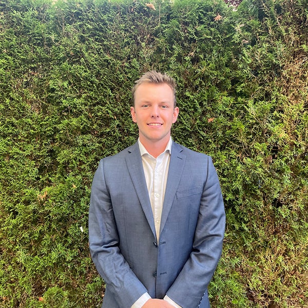 Max Dragon stands in front of a wall covered with greenery. He is wearing a grey suit and is smiling.