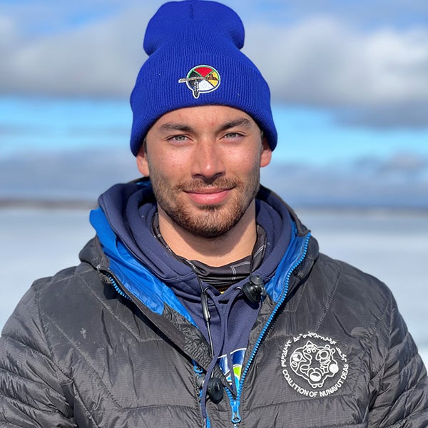 A picture of Qaritaq Kusugak-Clark looking at the camera with a slight grin. He is wearing a blue toque with a medicine wheel on it, and multiple layers with a light black puffy jacket