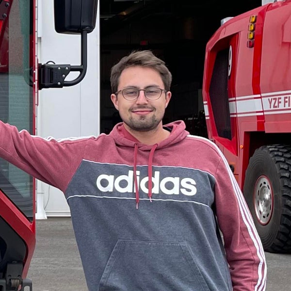 Scott Herman stands in front of what appears to be the firefighter bay at Yellowknife Airport. He has one hand outstretched and is leaning on a fire truck. He is wearing a maroon and grey adidas sweater, glasses, and is smiling at the camera.