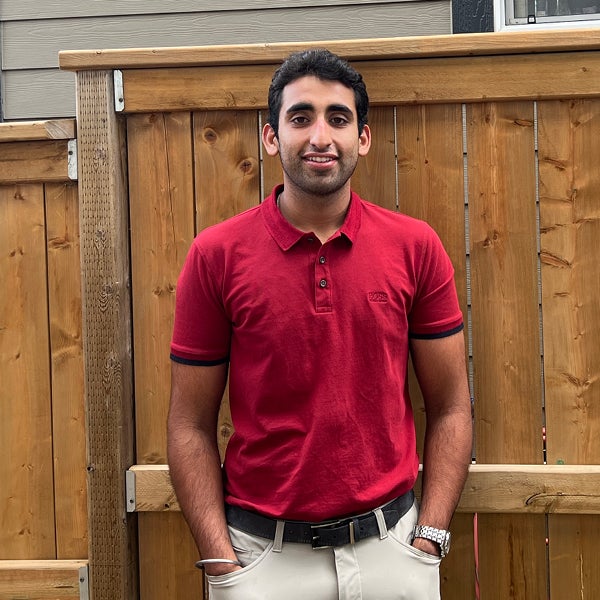 Viny Dhillon stands in front of a wooden fence wearing a red polo and white jeans. He is smiling.