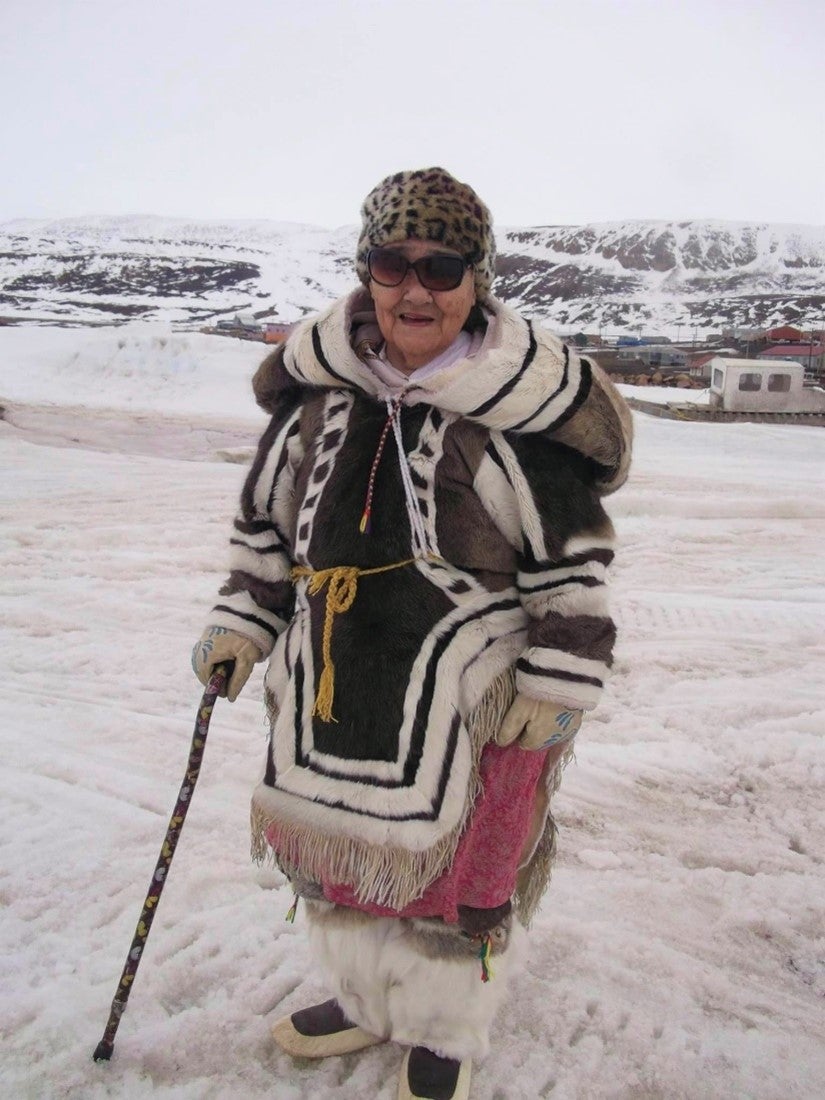 Kataisee's mother, Qapik Attagutsiak standing in traditional clothing she made. The background is a snowy landscape.