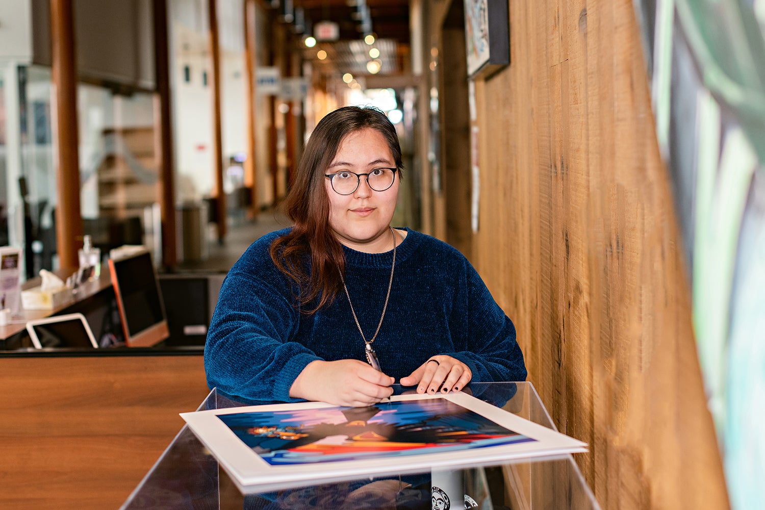 Kayla signing a lithograph of the Crow and The Native Spirit at the Kwanlin Dun Cultural Centre in Whitehorse.. They are sitting down and looking at the camera. The hallway blurs in the background, but looks to be made predominantly of Wood walls, and large glass windows.
