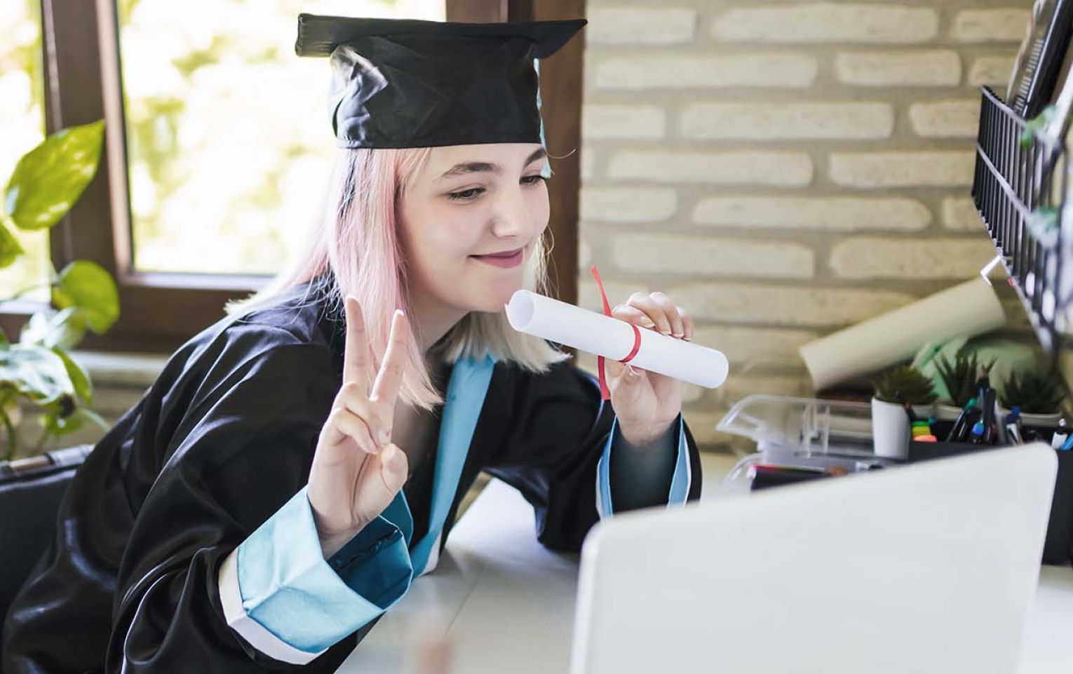 A young lady with light pink hair dressed in a graduation robe, sitting in front of a laptop. She is smiling, holding what appears to be a rolled up diploma. Her other hand is holding up her pointer and middle fingers - commonly referred to as the peace sign. In the background is a window lighting up the room.