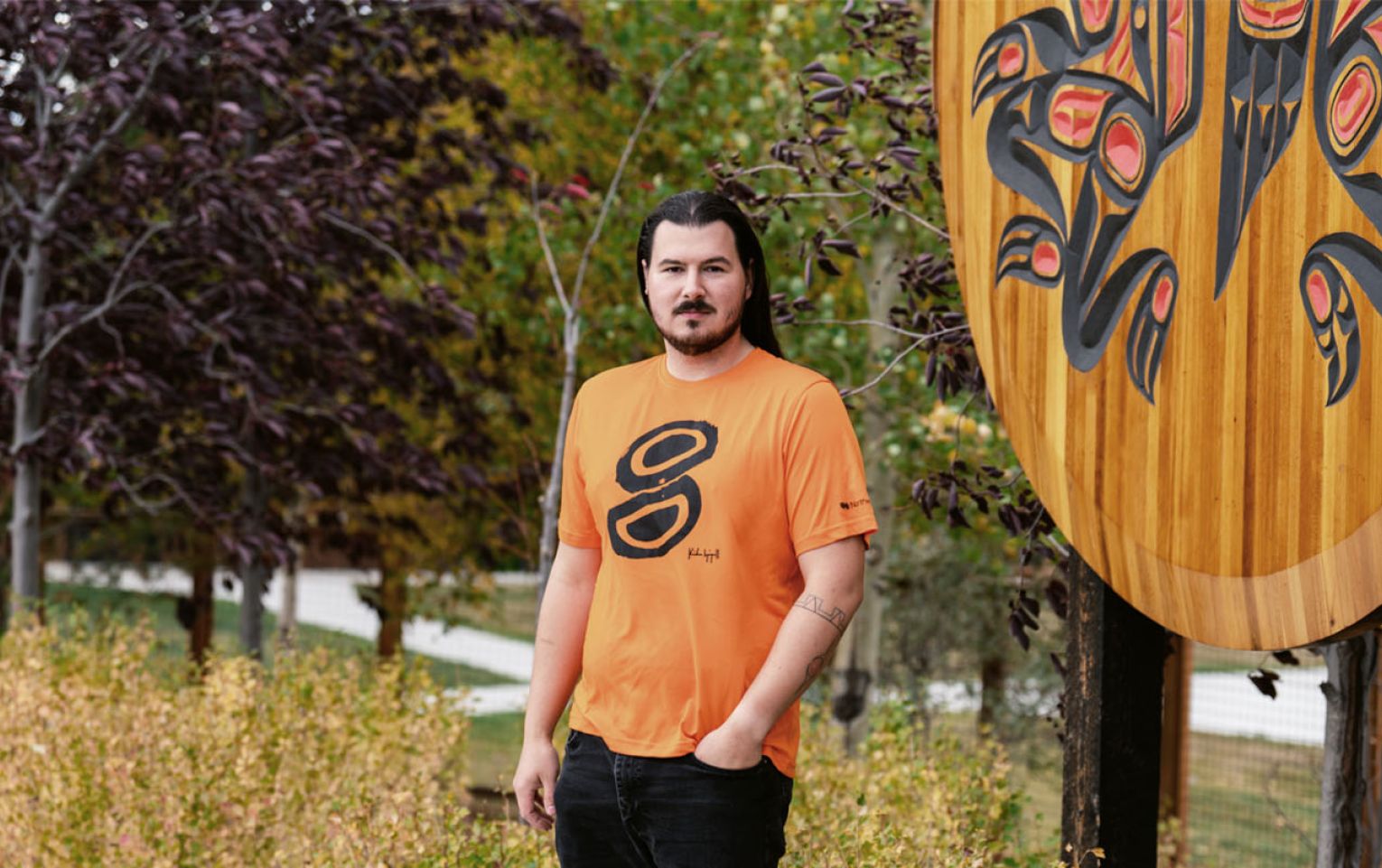 Kailen Gingall stands in front of a largen wooden crest at the Kwanlin Dun Cultural Centre. He is wearing the orange shirt he designed.