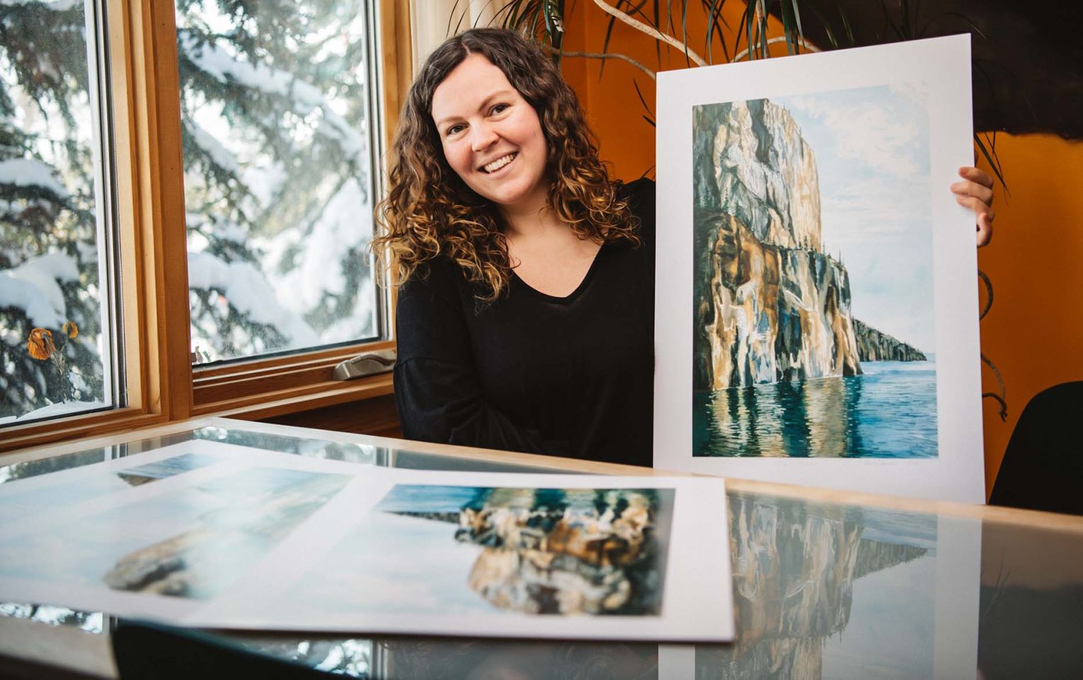 Maggie Davies, Northwest Territories Directory Art Contest winner, sits at a desk and smiles while holding a print of her art.  She is next to a window and snowy trees are visible outside. Inside, the room has a nice warm hue.