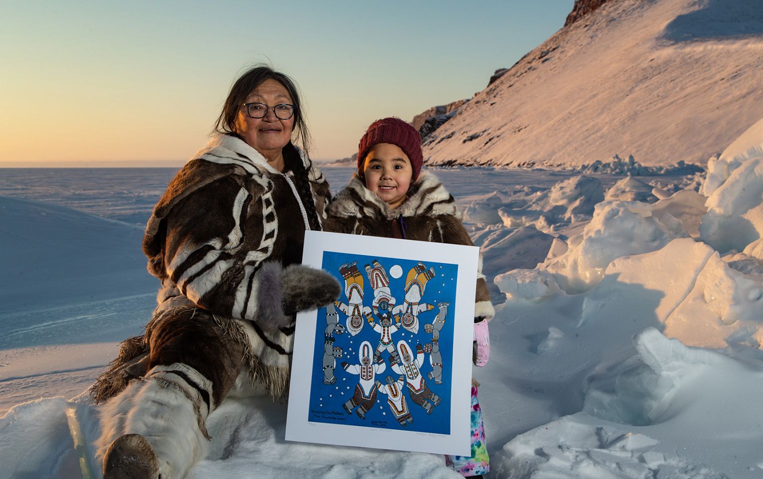 The winner of Nunavut's directory art contest,  Kataisee Attagutsiak with her young great granddaughter Nala holding a lithograph of the winning art. They are sending on the edge of a snowy, vast landscape. To the right, a snowy cliff shoots out of the earth at about at 45 degree angle. Both are wearing entirely traditional Inuit clothing which looks to be made of sealskin, and possibly caribou. Both are smiling widely.