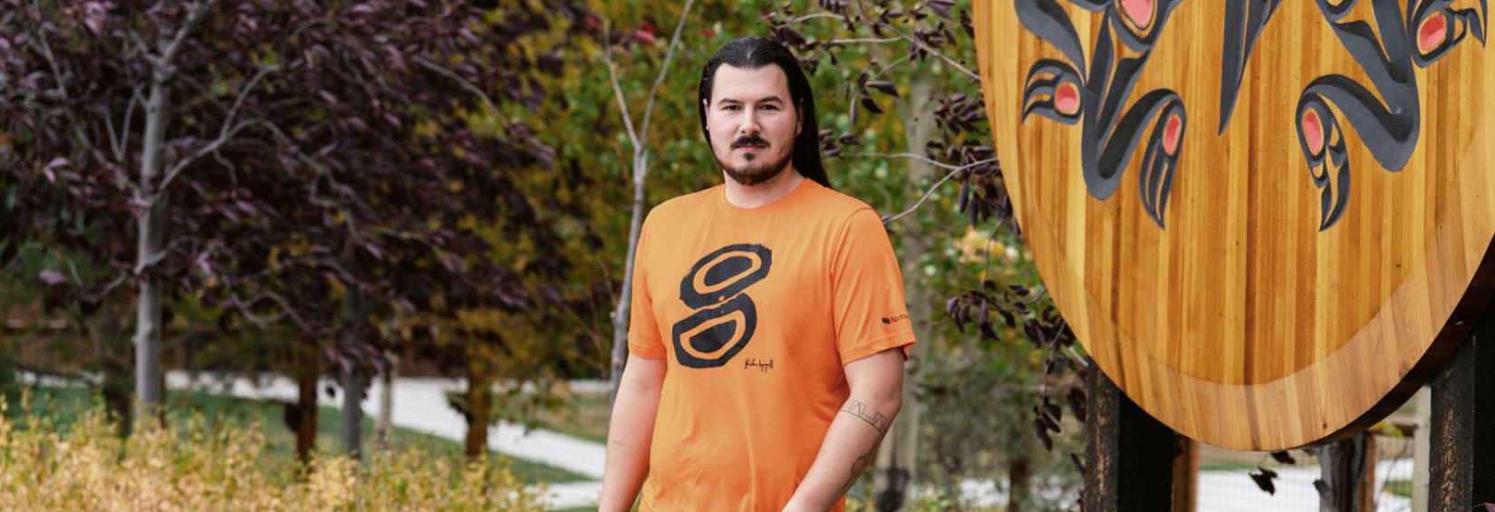Kailen Gingall stands in front of a largen wooden crest at the Kwanlin Dun Cultural Centre. He is wearing the orange shirt he designed.