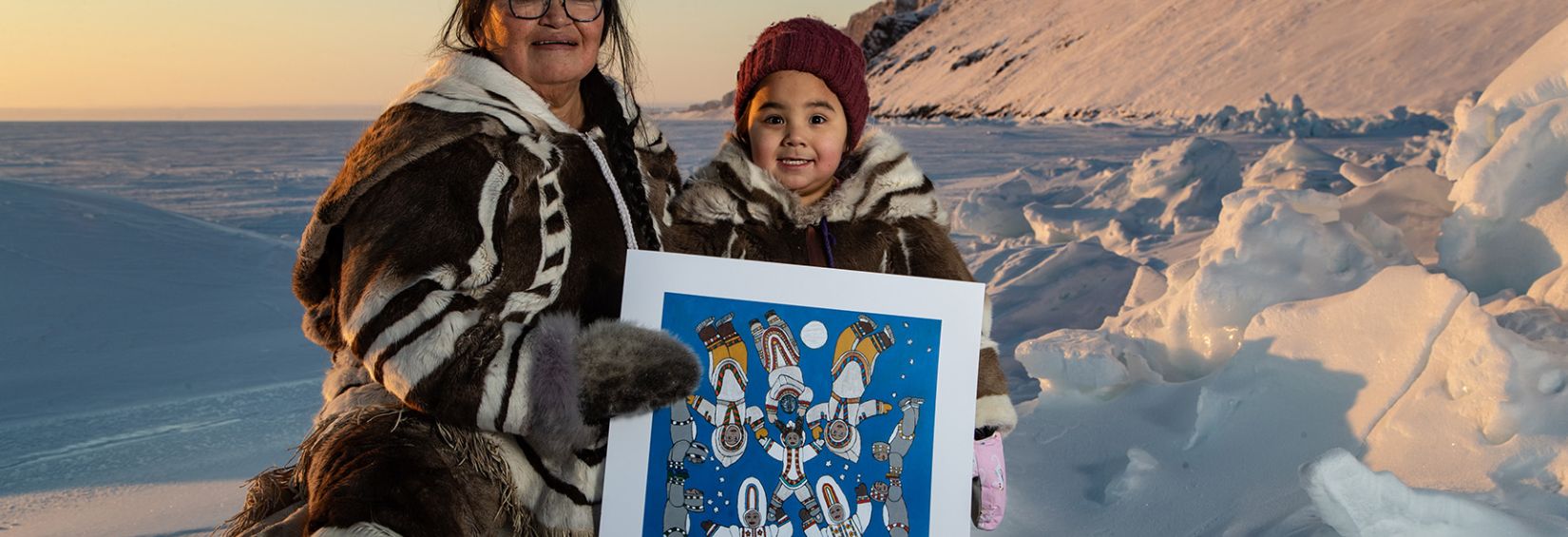 Kataisee and her great granddaughter Nala in Arctic Bay, Nunavut The winner of Nunavut's directory art contest, Kataisee Attagutsiak with her young great granddaughter Nala holding a lithograph of the winning art. They are sending on the edge of a snowy, vast landscape. To the right, a snowy cliff shoots out of the earth at about at 45 degree angle. Both are wearing entirely traditional Inuit clothing which looks to be made of sealskin, and possibly caribou. Both are smiling widely.