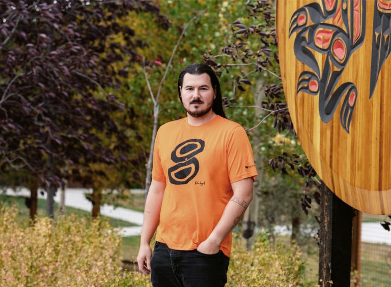 Kailen Gingall stands in front of a largen wooden crest at the Kwanlin Dun Cultural Centre. He is wearing the orange shirt he designed.